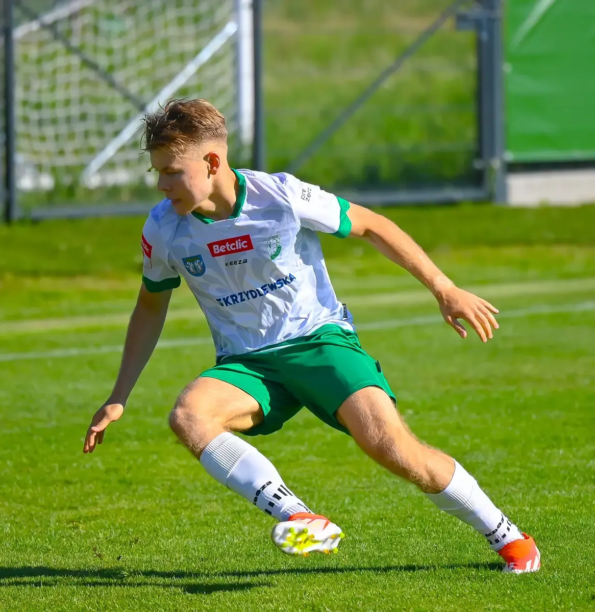 Zawodnik w ruchu Piłkarz Pelikana Łowicz w białej koszulce i zielonych spodenkach wykonuje krok na murawie obok bramki.