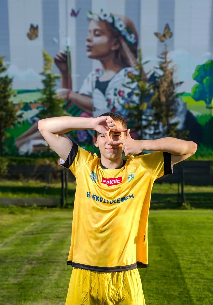Młodzież i wychowankowie Pelikana Młody zawodnik Pelikana Łowicz w żółtym stroju bramkarskim z logotypami sponsorów układa dłonie w kształt ramki na tle muralu.