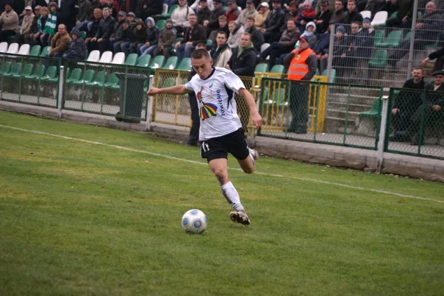 Zawodnik Pelikana Łowicz w białym stroju prowadzi piłkę podczas meczu przy stadionowych trybunach