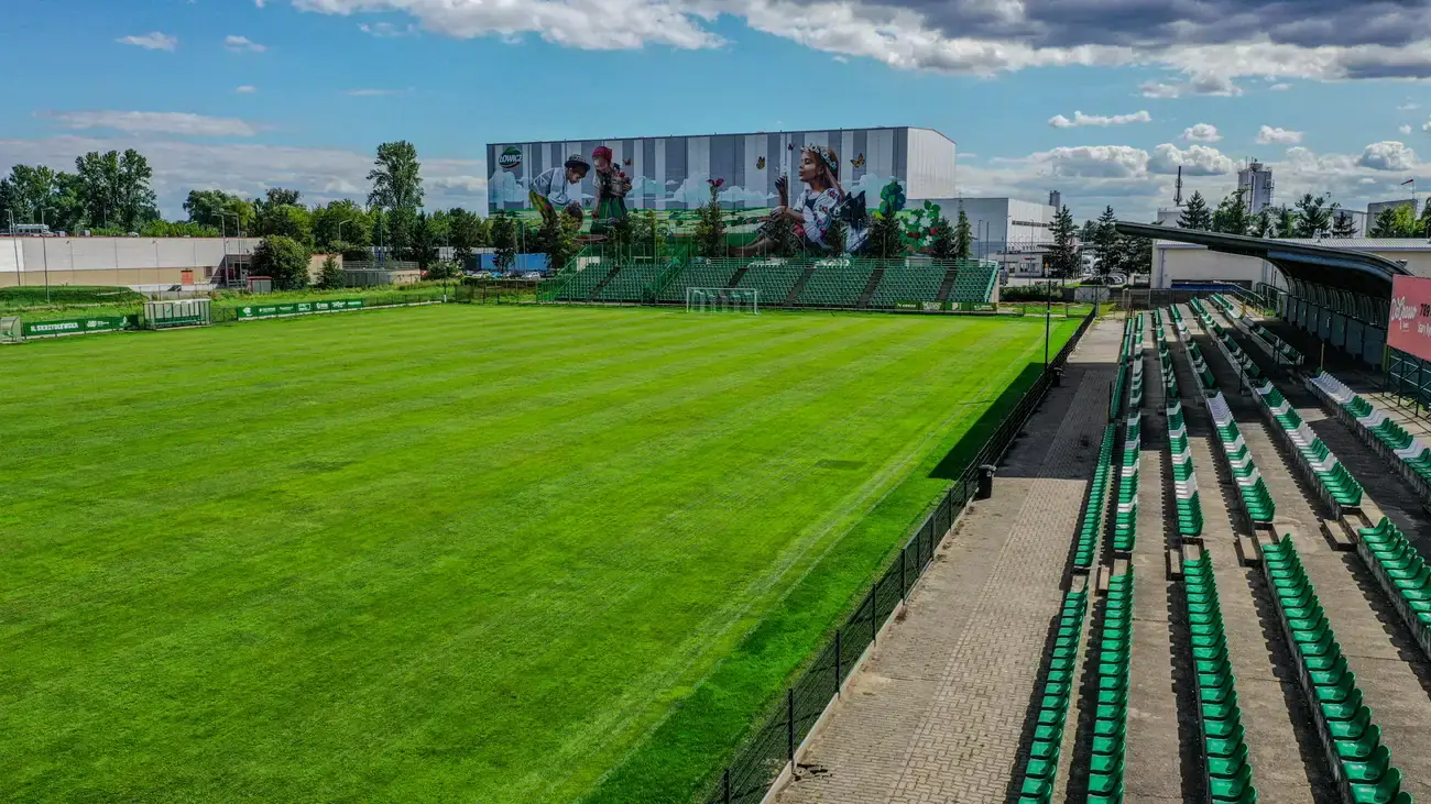 Widok z trybuny bocznej Stadionu Miejskiego w Łowiczu na murawę i zielone sektory z muralem w tle.