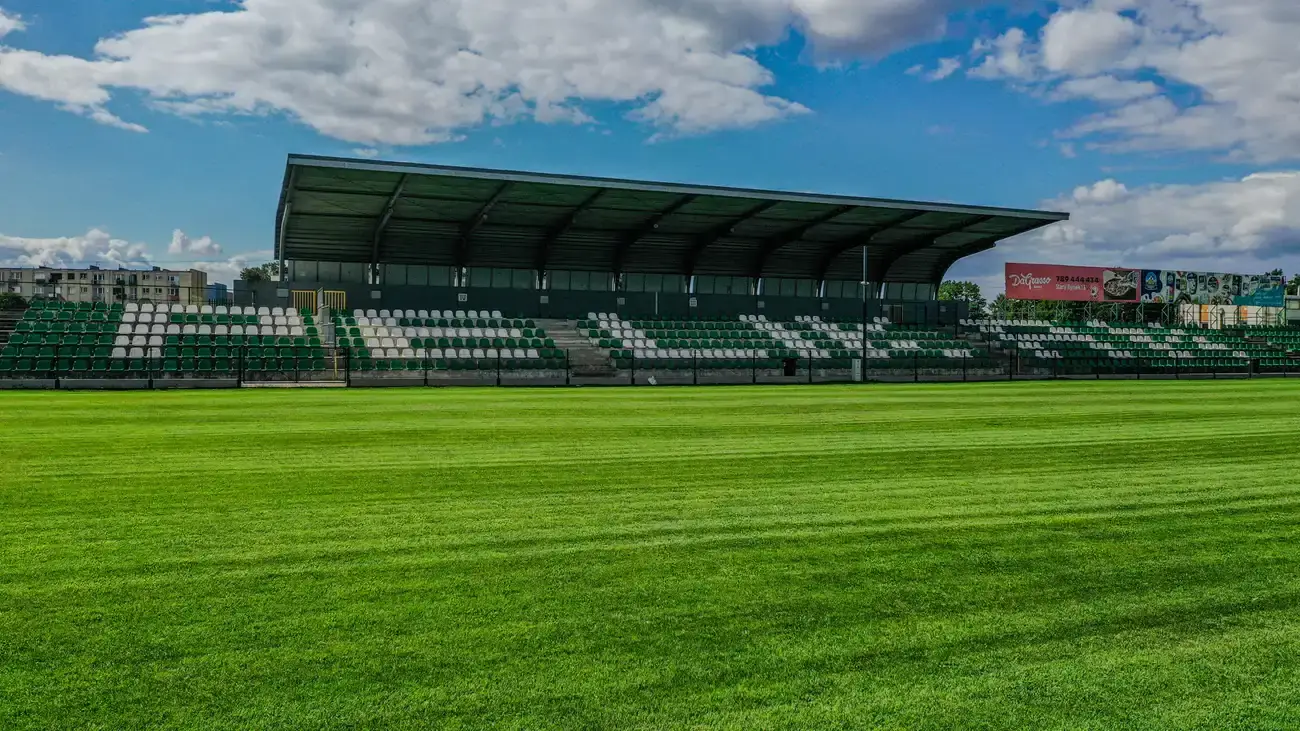 Zadaszona trybuna główna Stadionu Miejskiego w Łowiczu z biało-zielonymi krzesełkami i zadbaną murawą.