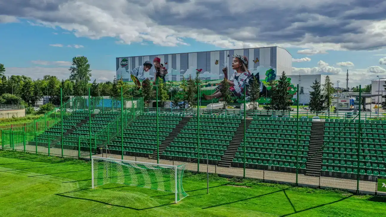 Zielone krzesełka trybuny bocznej Stadionu Miejskiego w Łowiczu z bramką treningową na pierwszym planie.