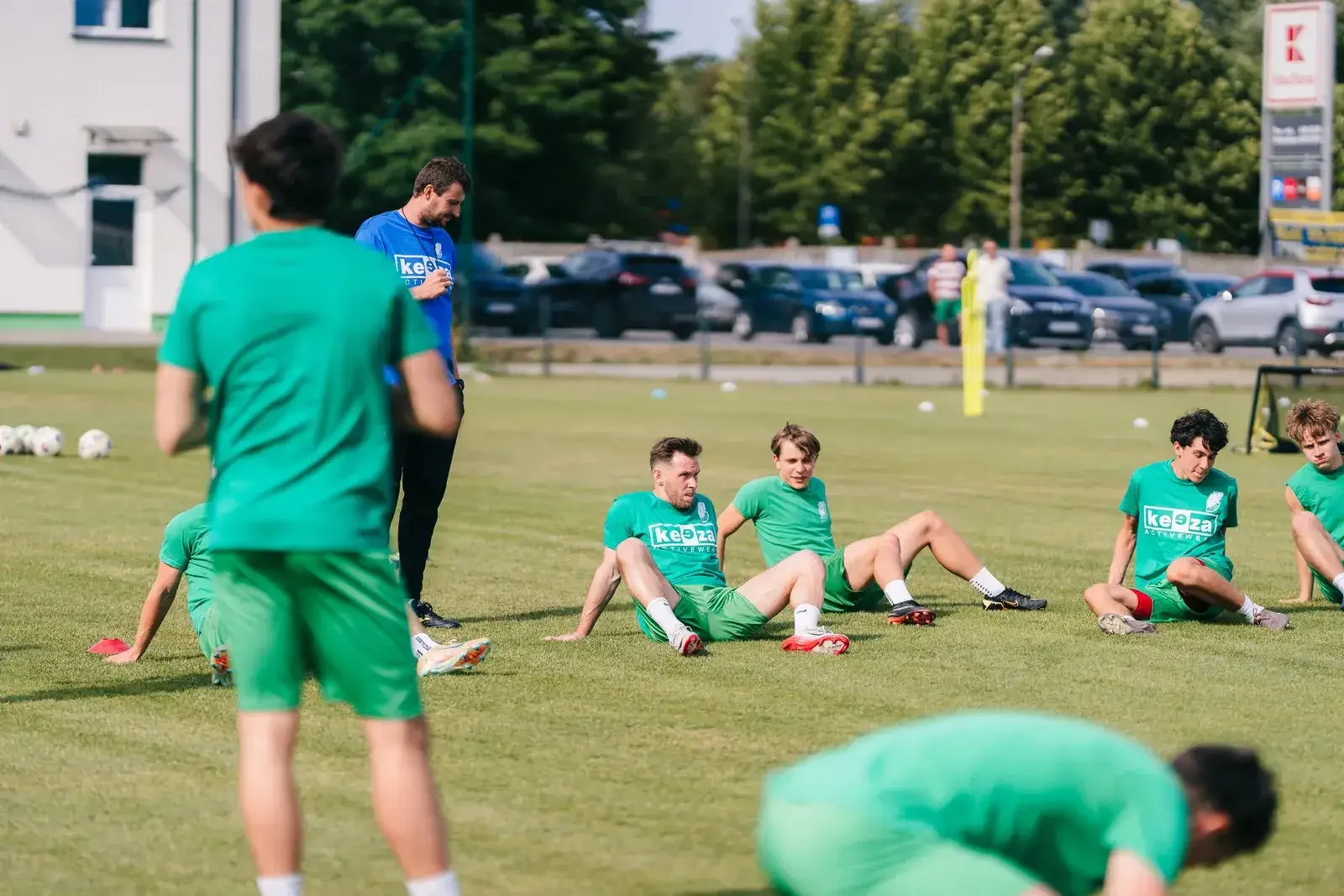 Ćwiczenia na siedząco Piłkarze Pelikana Łowicz siedzą na murawie podczas ćwiczeń, a osoba prowadząca trening stoi obok.
